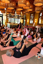 Group yoga class on pink mats in a warm indoor bar/event space with woven pendant lights, stocked back bar, mirrors and people stretching with legs raised.
