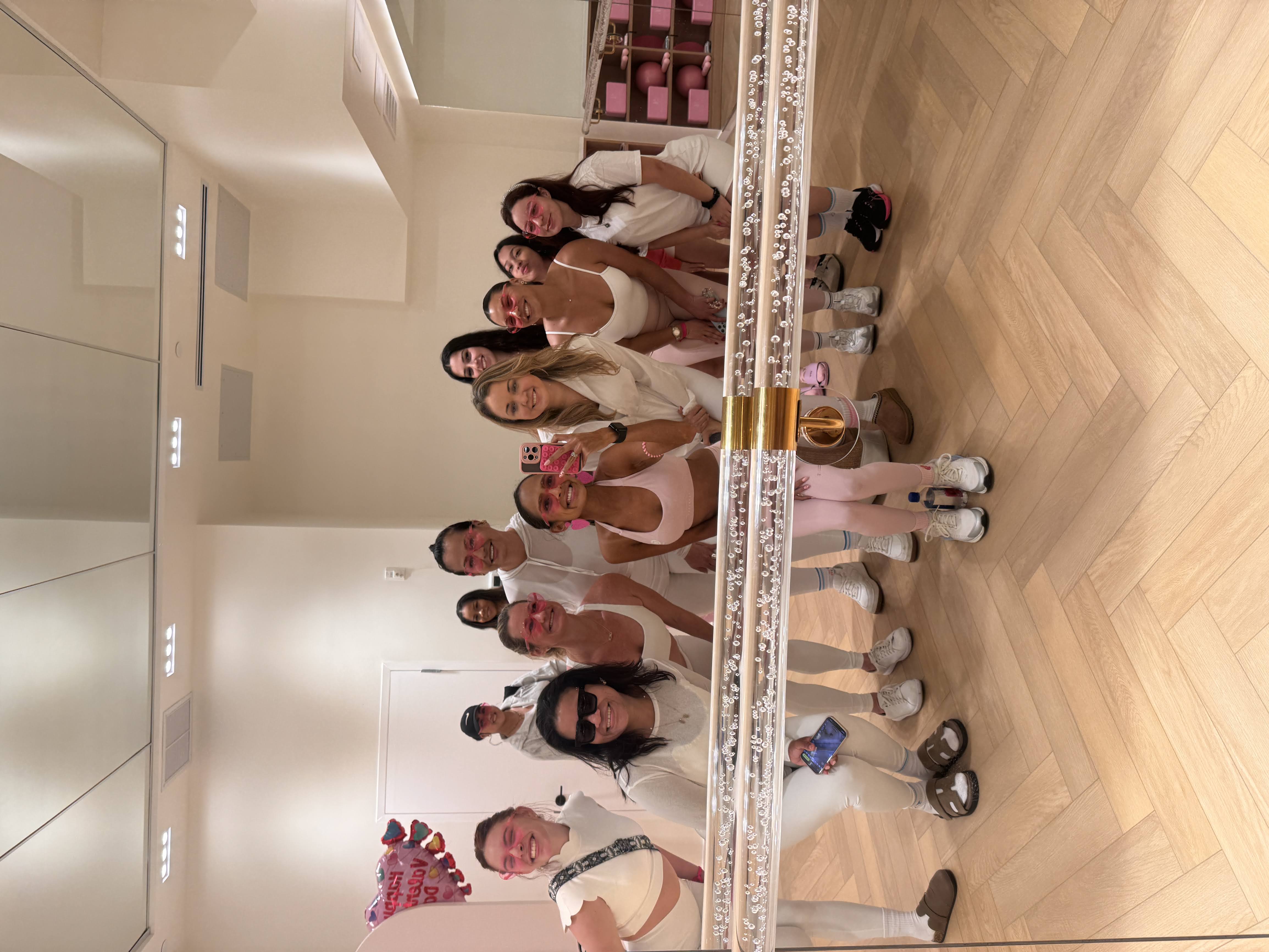 Group mirror selfie of smiling women in pink and white workout outfits inside a bright fitness studio with herringbone wood floor, mirrored wall and festive balloons