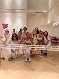 Group mirror selfie of smiling women in pink and white workout outfits inside a bright fitness studio with herringbone wood floor, mirrored wall and festive balloons