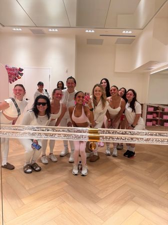 Group mirror selfie of smiling women in pink and white workout outfits inside a bright fitness studio with herringbone wood floor, mirrored wall and festive balloons
