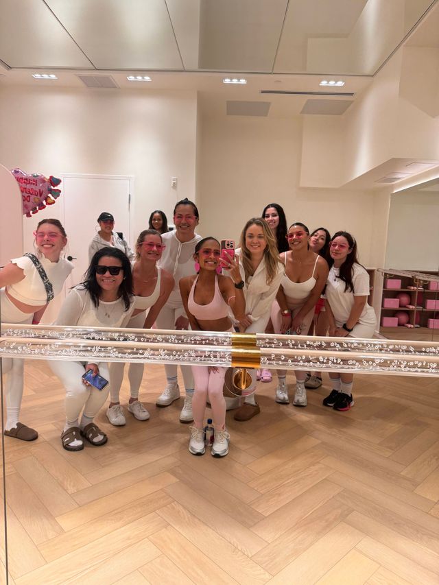 Group mirror selfie of smiling women in pink and white workout outfits inside a bright fitness studio with herringbone wood floor, mirrored wall and festive balloons