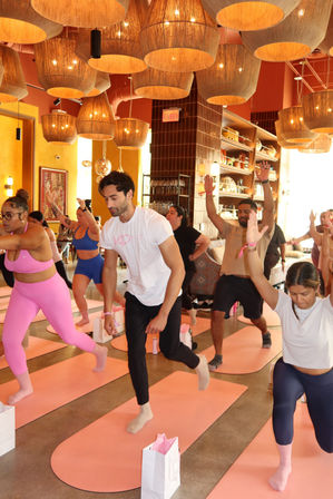 Boutique indoor yoga class in a bright wellness studio with pink mats and woven pendant lights, diverse participants practicing lunges and raised-arm poses.