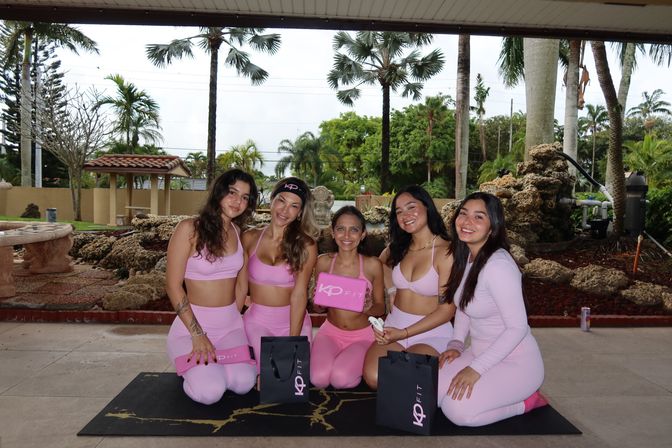 Five smiling women in matching pink activewear kneeling on a yoga mat under a covered tropical patio with palm trees, holding pink gift boxes and black gift bags, group fitness vibe