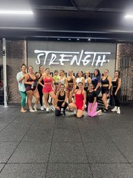 Smiling women in colorful activewear and one child pose for a group photo in a strength-training gym in front of a bold "STRENGTH" wall sign.