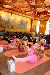 Group indoor yoga class on pink mats in a cozy yellow-walled studio with woven pendant lights and a vibrant wall mural; participants in colorful activewear doing supine knee-to-chest stretches.