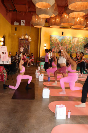 Group indoor yoga class in a warmly lit studio — participants in bright pink activewear practicing lunge poses on pink mats under large woven pendant lights with colorful wall art in the background.