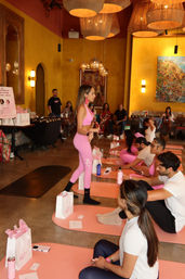 Instructor in bright pink activewear leading a group yoga and wellness workshop on pink mats inside a warm, lantern-lit boutique studio, with participants seated nearby holding water bottles and gift bags.