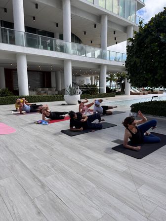 Outdoor poolside yoga class on mats at a modern beachfront building — white columns, glass balconies, palm trees and lounge chairs in the background.