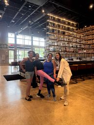 Four smiling friends in athletic clothing posing inside a modern industrial-style bar with high ceilings, exposed ductwork, large windows and a tall backlit shelving wall filled with bottles and glassware; one person lifts a pink-leggings-clad leg in a playful pose.