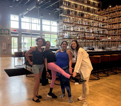 Four smiling friends in athletic clothing posing inside a modern industrial-style bar with high ceilings, exposed ductwork, large windows and a tall backlit shelving wall filled with bottles and glassware; one person lifts a pink-leggings-clad leg in a playful pose.