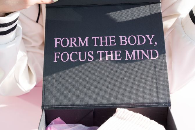 Black textured box lid with pink motivational text reading "FORM THE BODY, FOCUS THE MIND" resting on pastel pink clothing and gift packaging.