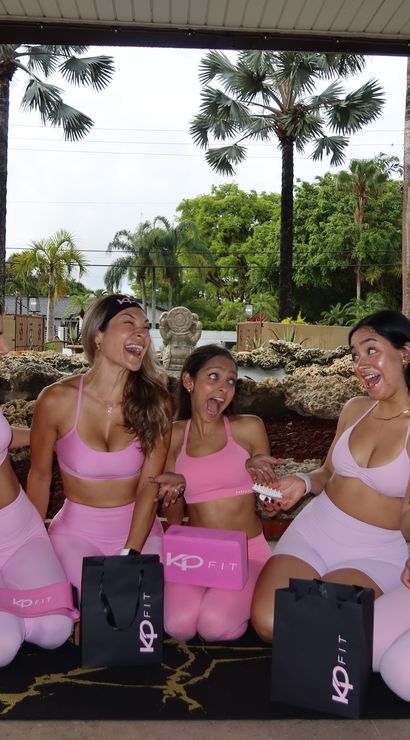Five women in matching pink workout sets laughing together on a covered tropical patio with palm trees, fitness mats and gift bags, outdoor group workout vibe
