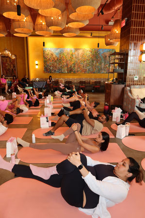 Community yoga class on pink mats in a warm, chandelier-lit studio — participants lie on their backs doing knee-to-chest stretches beneath woven pendant lights and a colorful mural.