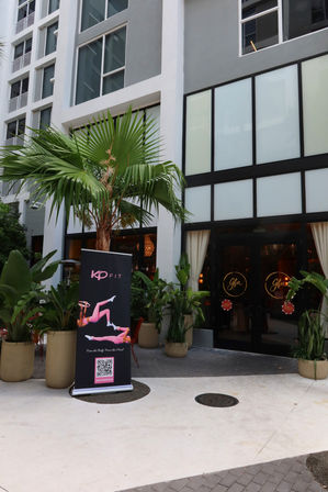 Modern urban storefront entrance with glass doors featuring circular gold logos, flanked by potted tropical plants and a small palm tree, and a black fitness promo banner with exercise silhouettes and a QR code on the sidewalk.