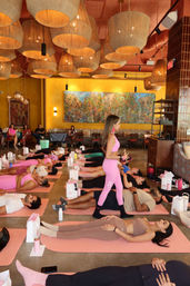 Group wellness class in a vibrant indoor studio — participants lying on pink mats during guided relaxation while an instructor in bright pink walks between them beneath oversized woven pendant lights and a colorful mural.