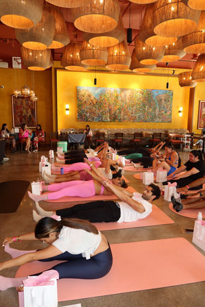 Group indoor yoga class on pink mats under woven pendant lights in a warm yellow studio with a large colorful mural