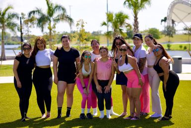 Smiling group of women in colorful activewear posing on a green lawn at a sunny waterfront park with palm trees — outdoor fitness meetup.