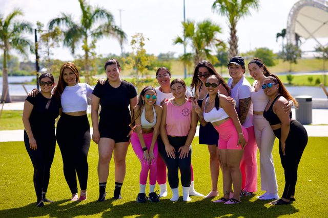 Smiling group of women in colorful activewear posing on a green lawn at a sunny waterfront park with palm trees — outdoor fitness meetup.