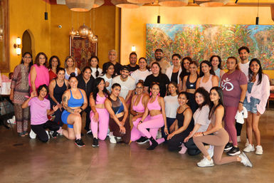 Large smiling group of adults at a fitness and wellness meetup, many in pink activewear, posing indoors in a warm-lit room with hanging lanterns and a colorful mural — community group photo.