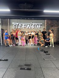 Group of men and women in colorful workout clothes posing in a gym in front of a large “STRENGTH” mural, with mats and fitness equipment on the floor
