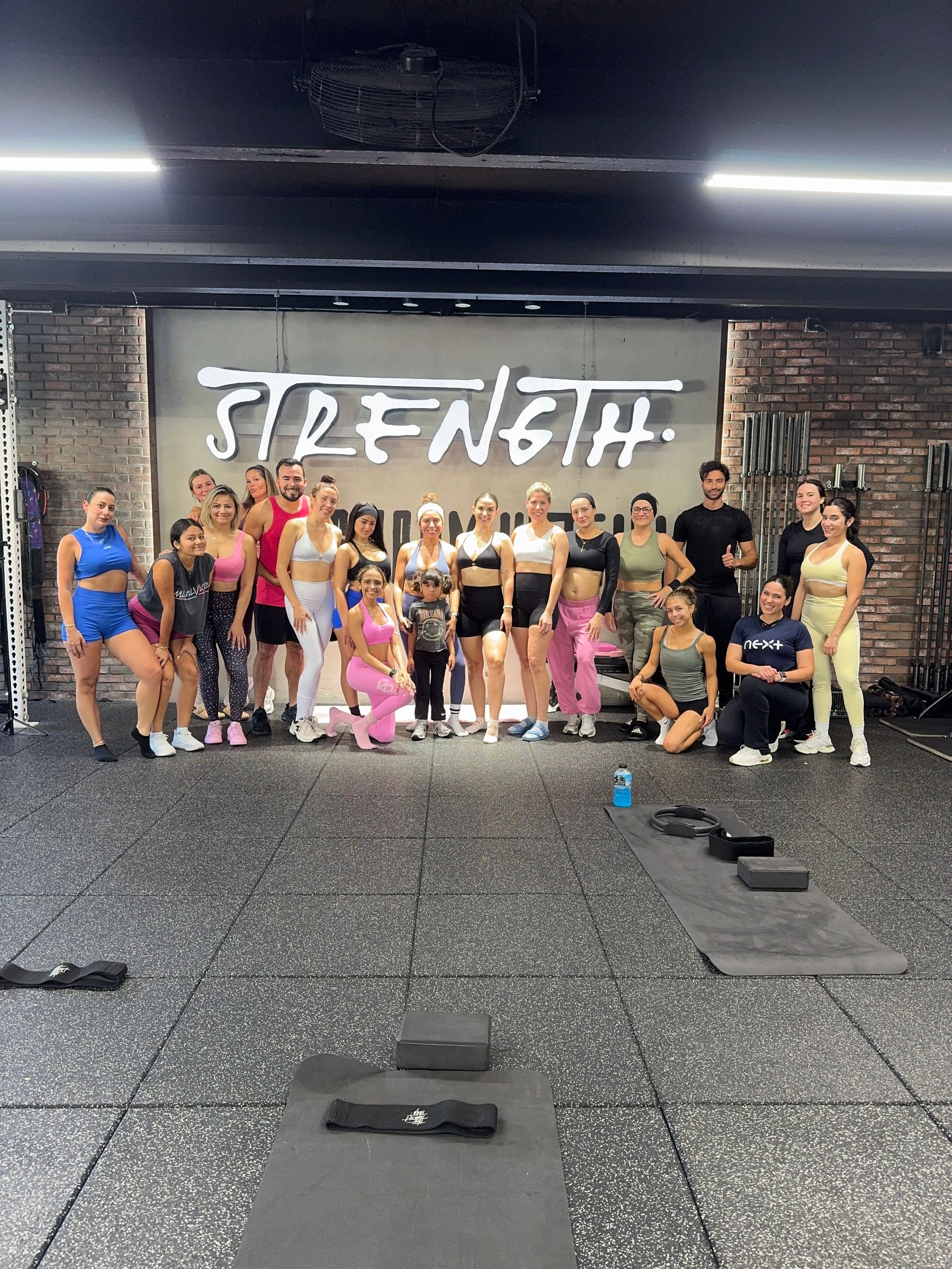 Group of men and women in colorful workout clothes posing in a gym in front of a large “STRENGTH” mural, with mats and fitness equipment on the floor
