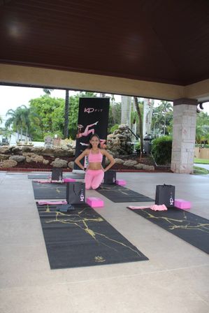 Woman in pink activewear kneeling on a yoga mat in a covered outdoor pavilion with palm trees, black-and-gold mats and pink yoga props set up for a tropical fitness class