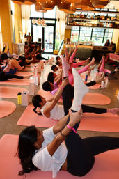 Energetic indoor yoga session on pink mats in a bright wellness studio — participants lying on their backs holding raised legs during a guided stretch, warm lighting and a bar-area backdrop.