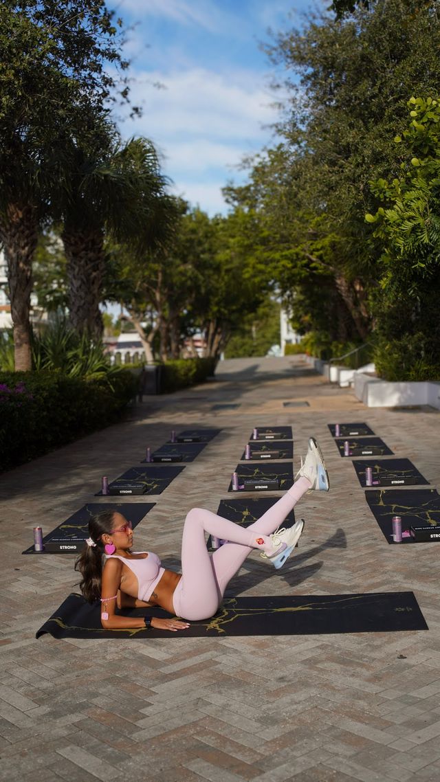 Outdoor yoga/fitness class on a sunny, palm-lined promenade — woman in pink activewear doing a floor leg lift on a black mat with rows of mats and water bottles ready for a group workout.