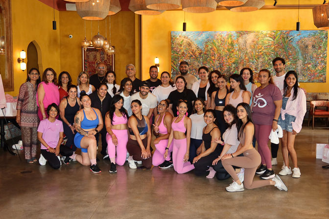 Group of men and women posing after a yoga and wellness workshop in a bright indoor studio, many wearing pink activewear, warm yellow walls and a colorful mural.