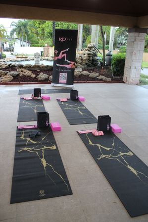 Outdoor yoga class setup on a covered patio with black mats featuring gold veining, pink yoga blocks, straps and small gift bags arranged in rows, tropical palms and rock landscaping visible in the background.
