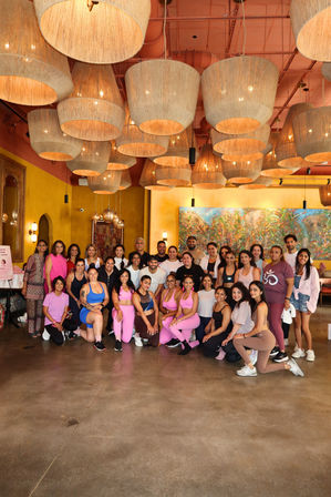 Smiling group of fitness participants posing in a vibrant indoor studio after a workout — warm yellow walls, oversized woven pendant lights and a colorful mural in the background.