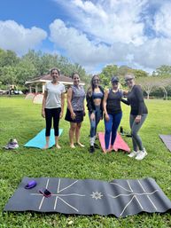 Five women posing on yoga mats during an outdoor yoga class in a neighborhood park with a gazebo, playground, green lawn and blue sky with puffy clouds