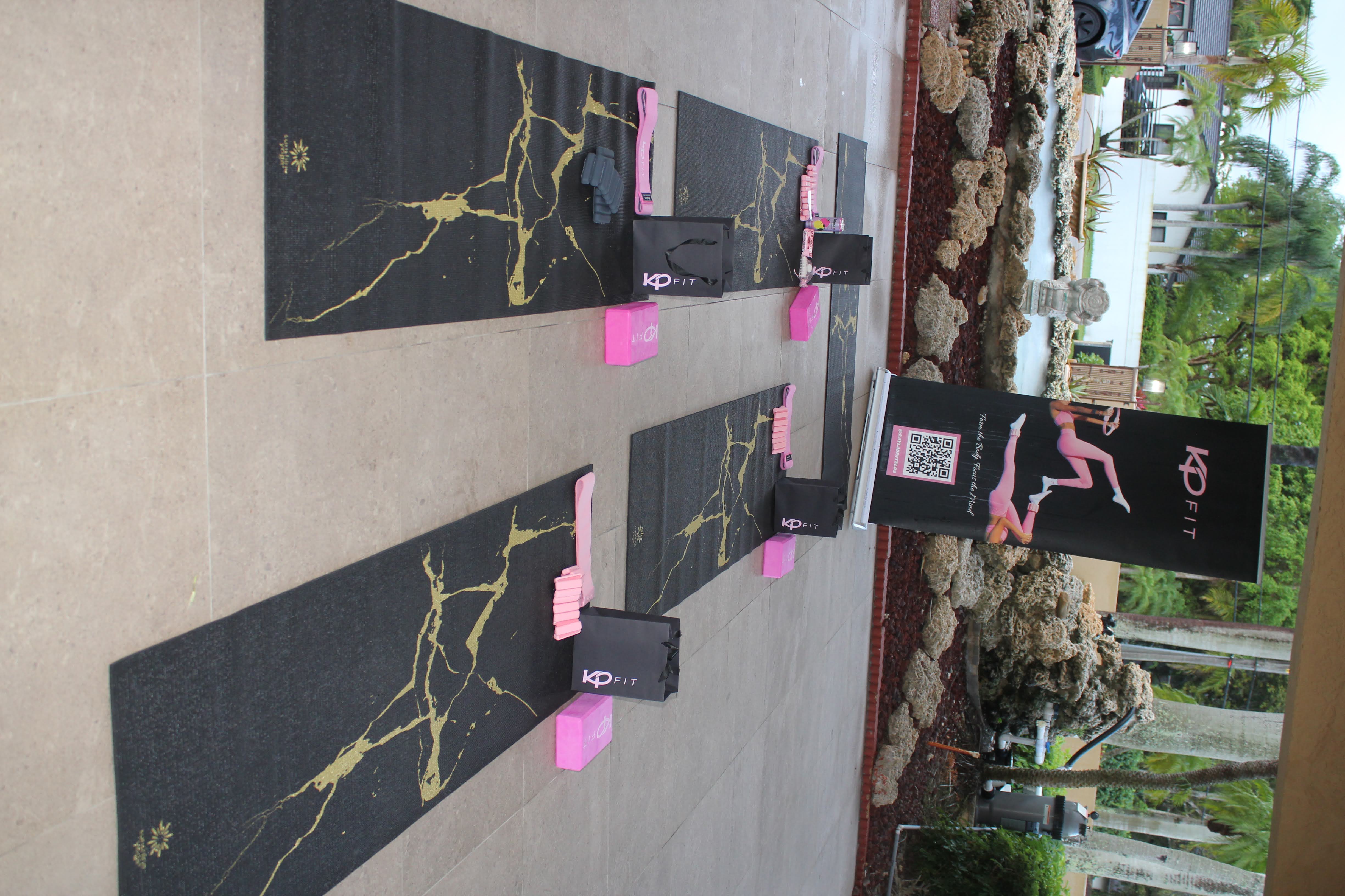 Outdoor tropical yoga class setup on a covered patio with black-and-gold mats, pink blocks, resistance bands and gift bags arranged near a rock fountain and palm trees