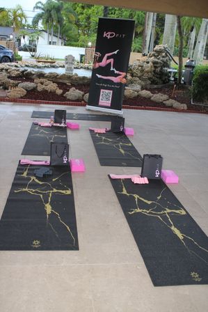 Outdoor tropical yoga class setup on a covered patio with black-and-gold mats, pink blocks, resistance bands and gift bags arranged near a rock fountain and palm trees