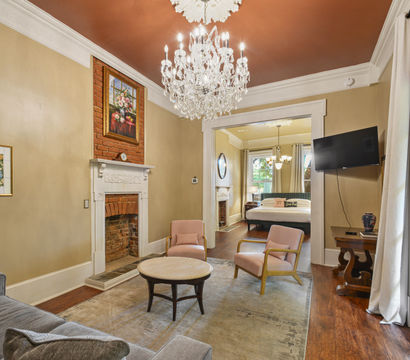 Period-style living room with a sparkling crystal chandelier, exposed brick fireplace with white mantel, blush armchairs and round coffee table, gray sofa on hardwood floors, French doors with curtains and an open view into a sunlit bedroom.