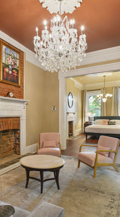 Period-style living room with a sparkling crystal chandelier, exposed brick fireplace with white mantel, blush armchairs and round coffee table, gray sofa on hardwood floors, French doors with curtains and an open view into a sunlit bedroom.