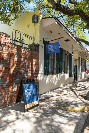 Sunlit sidewalk outside a small boutique hotel in New Orleans with a blue hanging sign and A-frame board by a brick wall, green shutters and dappled oak tree shadows.