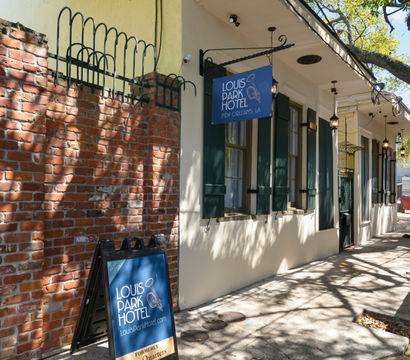 Sunlit sidewalk outside a small boutique hotel in New Orleans with a blue hanging sign and A-frame board by a brick wall, green shutters and dappled oak tree shadows.
