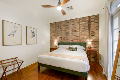 Bright cozy bedroom with exposed brick accent wall, green upholstered bed, hardwood floors, ceiling fan, bedside lamps and framed bird artwork
