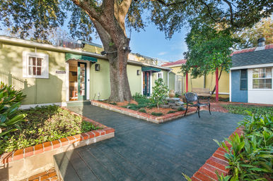 Cozy sunny courtyard patio with a large oak tree, brick-edged planting beds, dark wooden deck, metal bench and wooden swing outside pastel green and yellow cottage-style buildings.