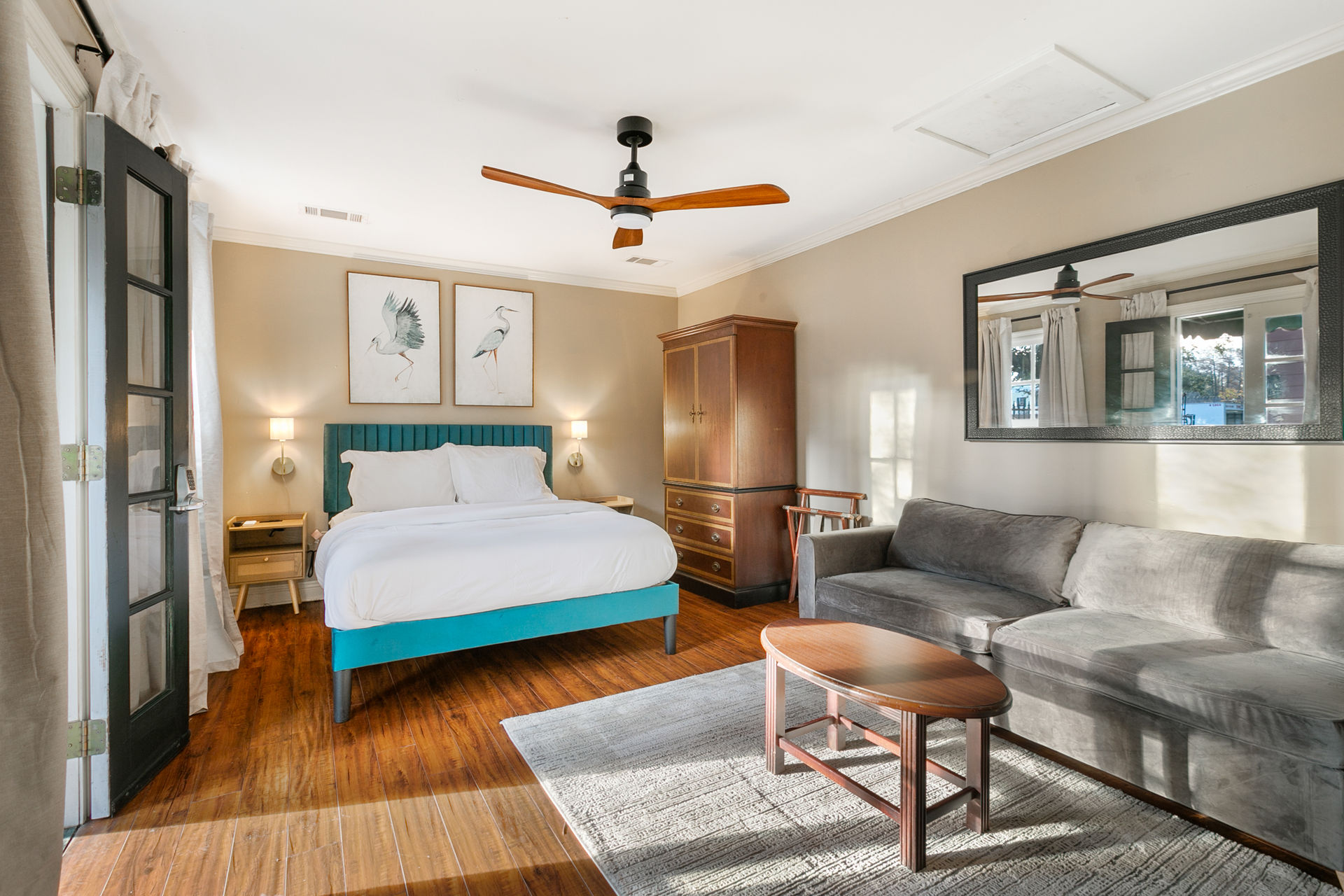 Cozy sunlit bedroom with teal-upholstered bed and white linens, wooden ceiling fan, gray sofa, large wall mirror, wooden armoire and bedside lamps on warm hardwood floors.
