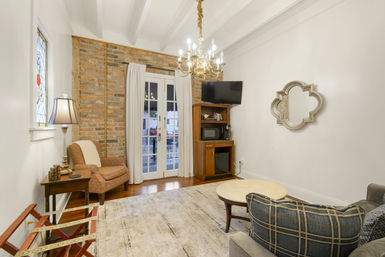 Cozy historic-style living room with exposed brick wall, French doors, gold chandelier, patterned armchair, wooden media cabinet with TV and microwave, decorative quatrefoil mirror, stained-glass window, neutral area rug and hardwood floors.