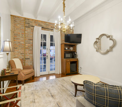 Cozy cottage living room with exposed brick accent wall, stained-glass window, French doors, brass chandelier, patterned armchair, TV cabinet and hardwood floors.