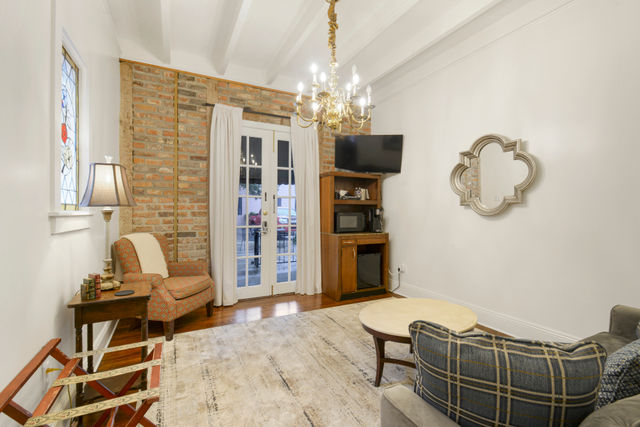 Cozy cottage living room with exposed brick accent wall, stained-glass window, French doors, brass chandelier, patterned armchair, TV cabinet and hardwood floors.