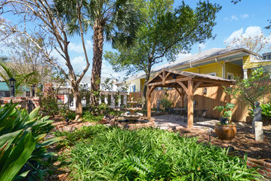 Sunny backyard garden patio with wooden pergola, stone fountain, outdoor seating and lush tropical plants near a yellow house