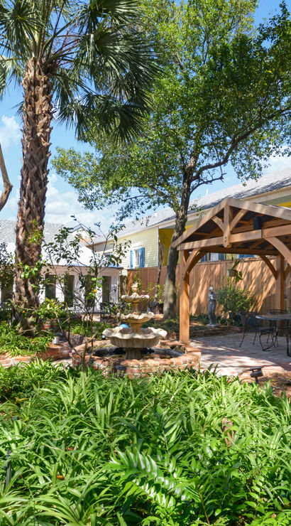 Sunny backyard garden patio with wooden pergola, stone fountain, outdoor seating and lush tropical plants near a yellow house