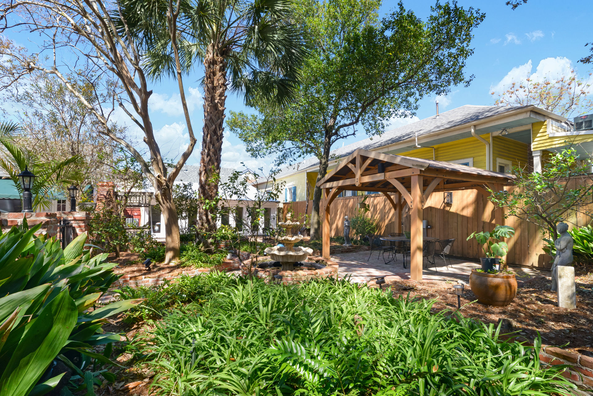 Sunny backyard garden patio with wooden pergola, stone fountain, outdoor seating and lush tropical plants near a yellow house