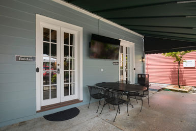Covered outdoor patio with light-blue siding and white French doors, wall-mounted TV, black metal dining table and chairs on a concrete floor, with a red building and small tree nearby.