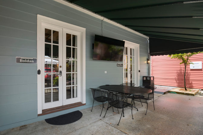 Covered outdoor patio with light-blue siding and white French doors, wall-mounted TV, black metal dining table and chairs on a concrete floor, with a red building and small tree nearby.