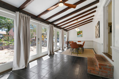 Cozy sunlit dining sunroom with exposed dark wood beams and ceiling fan, floor-to-ceiling French doors with light curtains opening to a brick patio, black tile floor, area rug and rust-colored dining chairs.